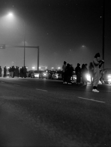 A late-night gathering of people and cars under streetlights in a foggy environment, with signs and 20km speed limit visible.