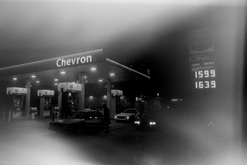 Chevron gas station at night with cars refueling; prices displayed are 159.9 and 163.9.