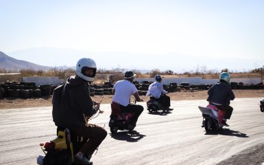 Group of people in helmets riding scooters on a sunny day, with desert landscape and tires in the background.