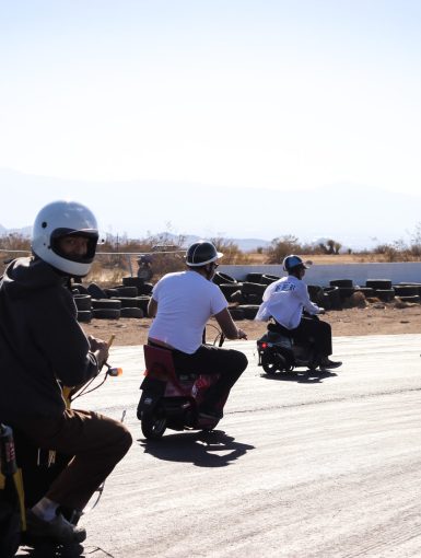 Group of people in helmets riding scooters on a sunny day, with desert landscape and tires in the background.