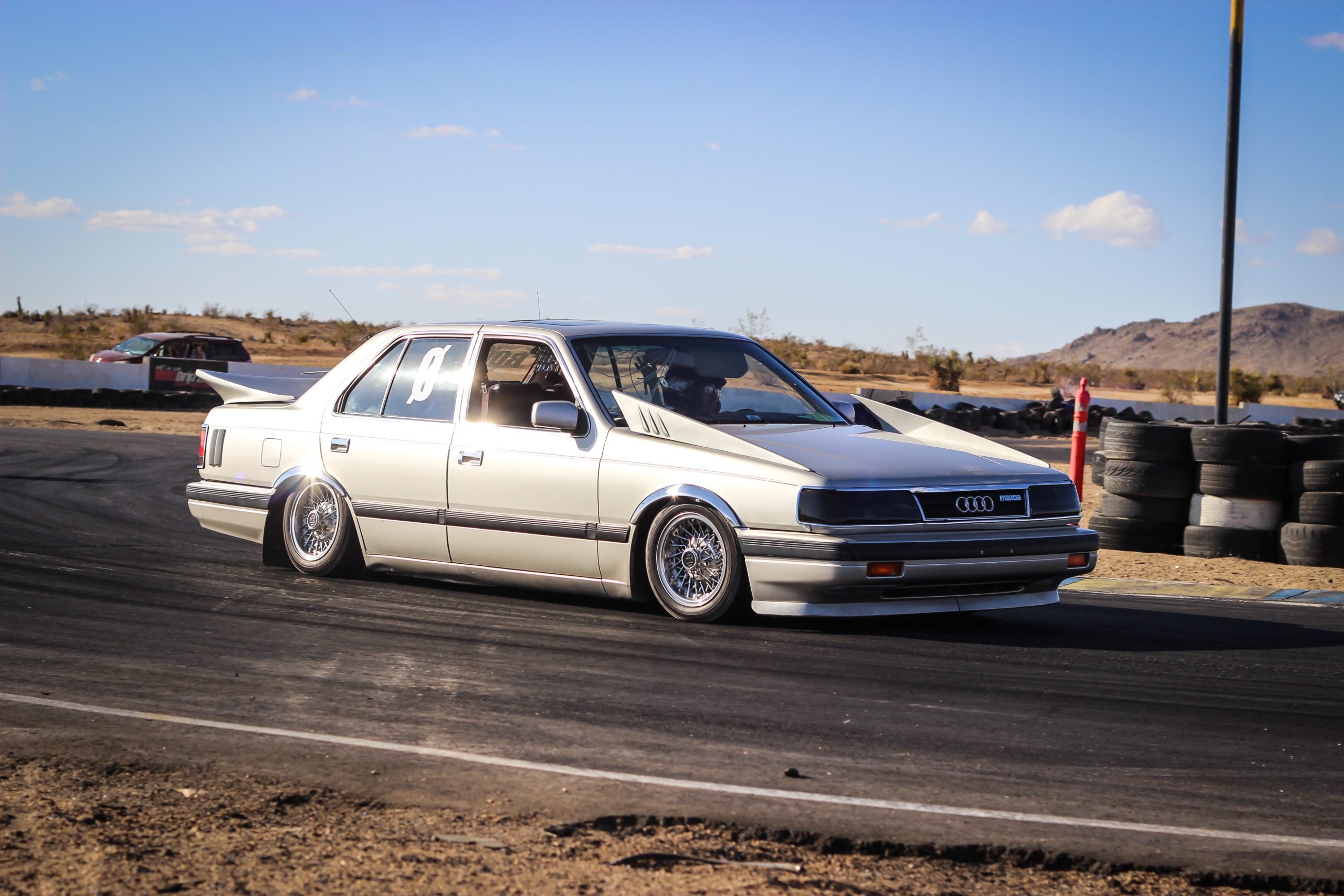 A modified white Audi sedan drifting on a race track with mountainous desert background and tire barriers.