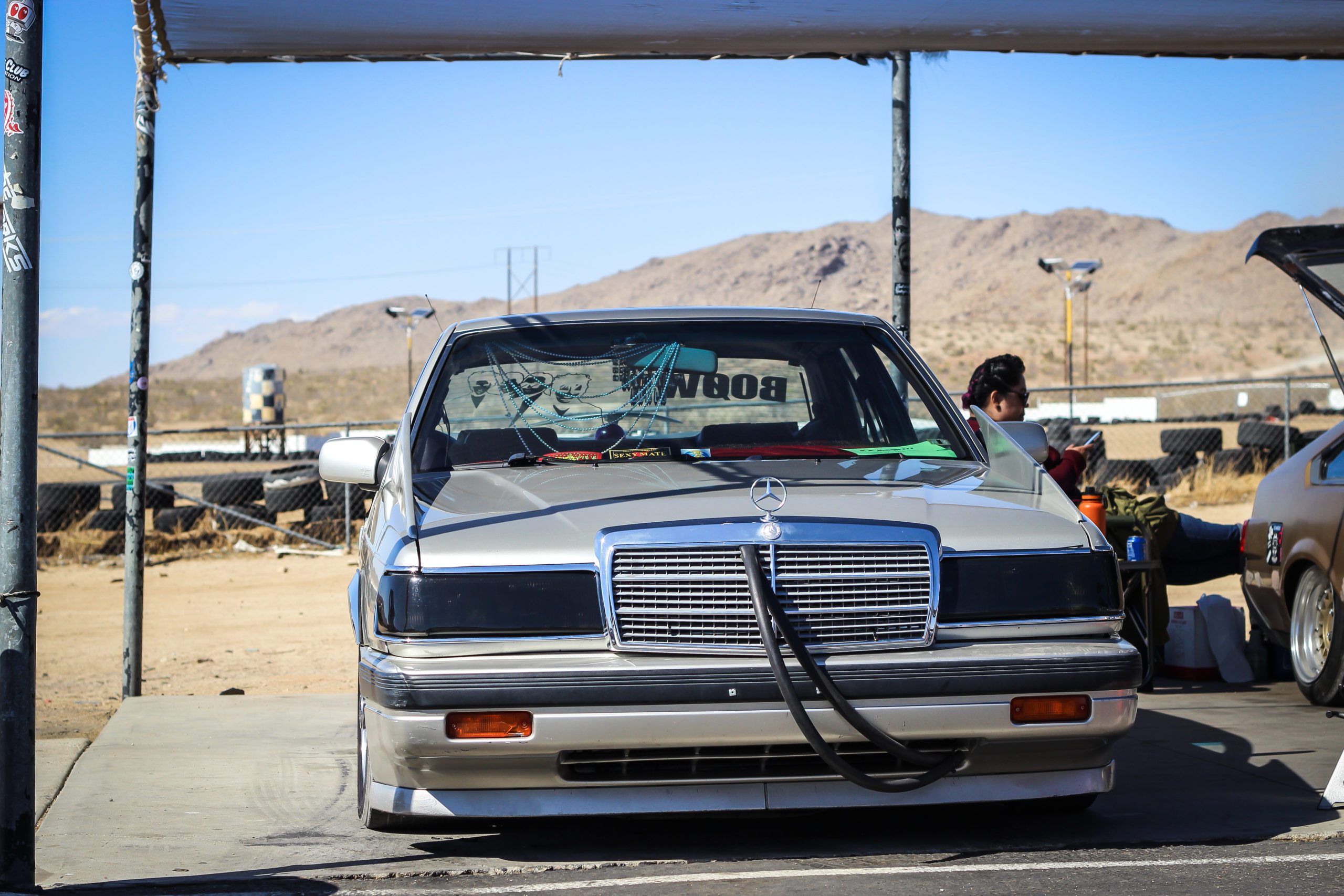 Mercedes-Benz parked under canopy at car event, with BOOM sticker in Japanese on rear windshield. Desert landscape in background.