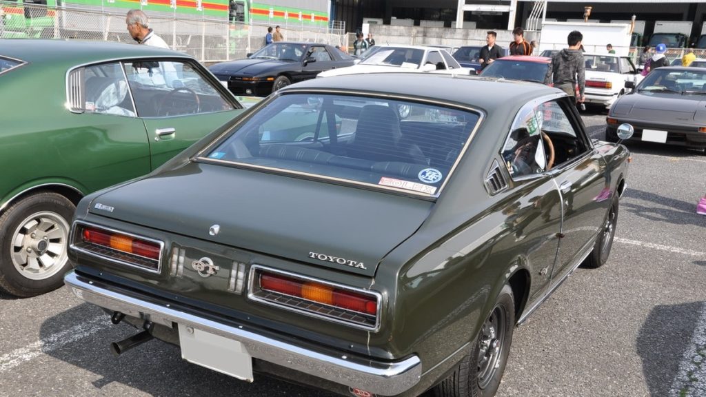Vintage Toyota GT on display at a car meet, surrounded by other classic cars; stickers in Japanese translate to SOLTEX.