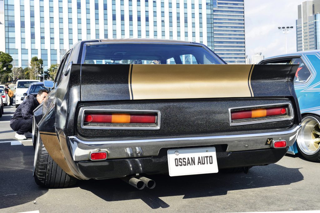 Vintage muscle car at a car show with OSSAN AUTO license plate. Buildings and people are visible in the background.