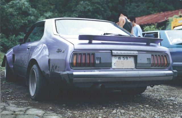 Alt text: Classic purple 1970s sports car with spoiler, Japanese license plate reading '59', parked on a gravel driveway.