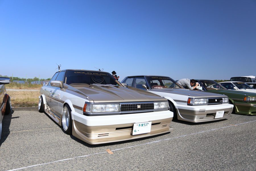 Modified vintage Japanese cars on display at a car meet, sunny day with blue skies. License plates in Japanese.