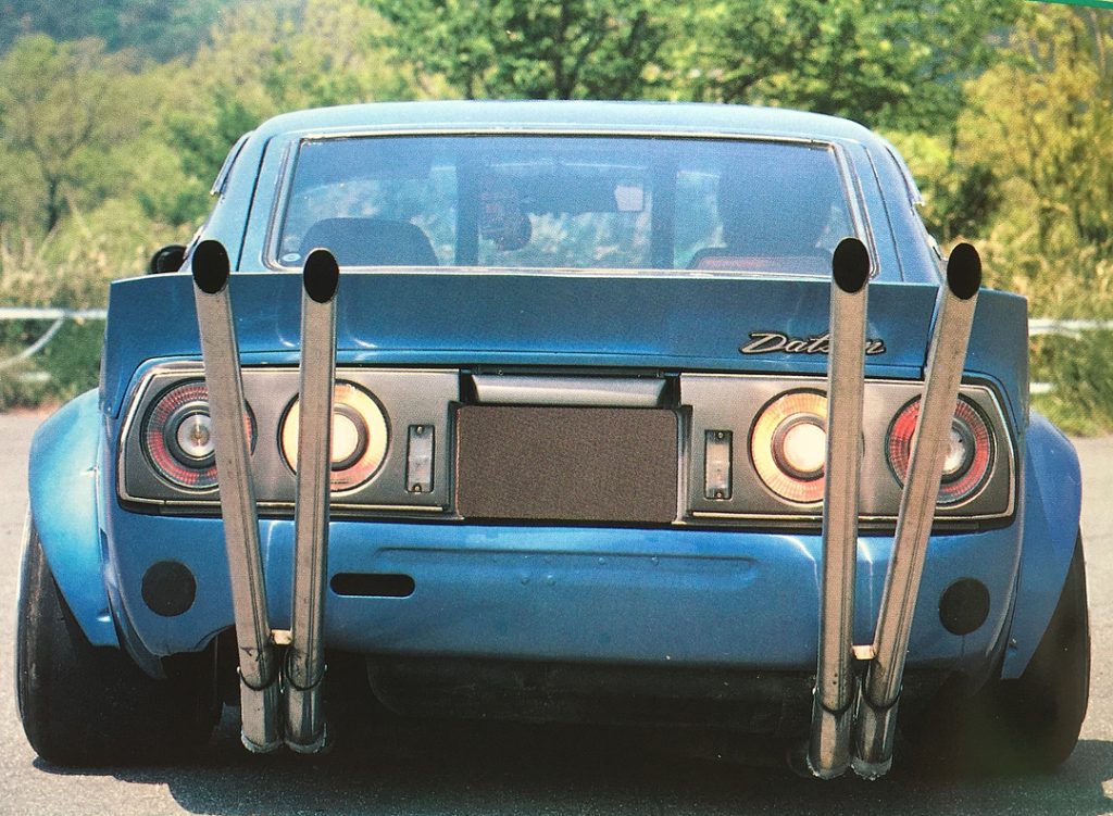 Rear view of a blue Datsun car with four tall exhaust pipes, showcasing a unique custom modification in a scenic outdoor setting.