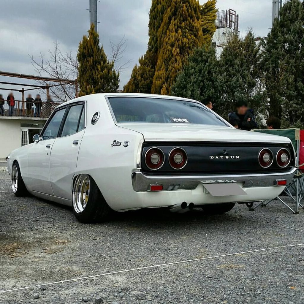 Retro white Datsun sedan with lowered suspension, displayed outdoors at a car show. Japanese text translates to Takumi.