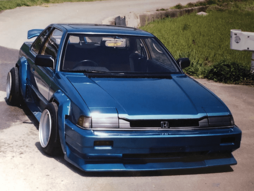 Modified blue Honda with wide body kit and TRUST sticker on windshield parked on a road.