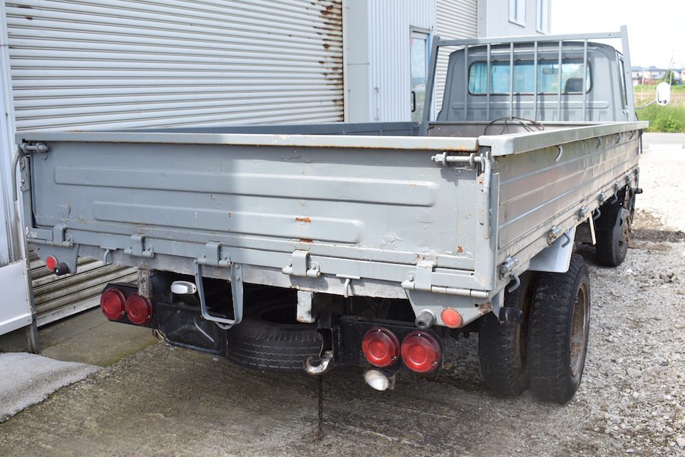 Rear view of an old, grey pickup truck parked near a building, showcasing its tailgate and taillights.