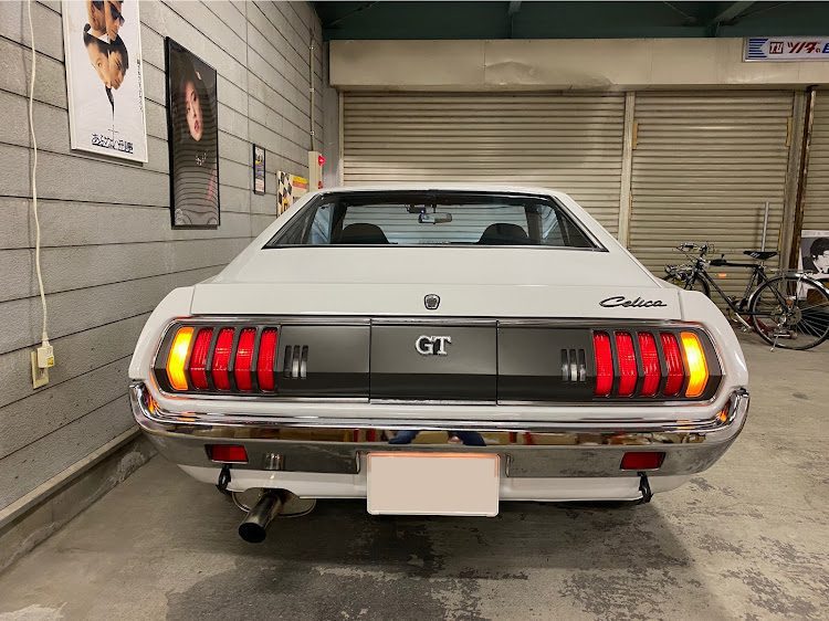 Alt text: Rear view of a classic white Toyota Celica GT in a garage with wall posters and bicycles in the background.