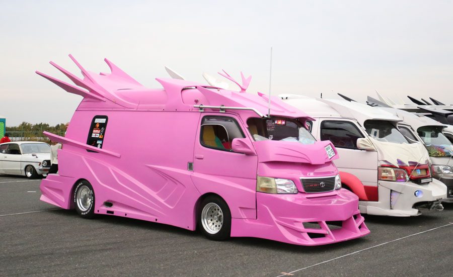 Pink custom vans with exaggerated body modifications at a car show in Japan. The pink van has Japanese decals on the side.