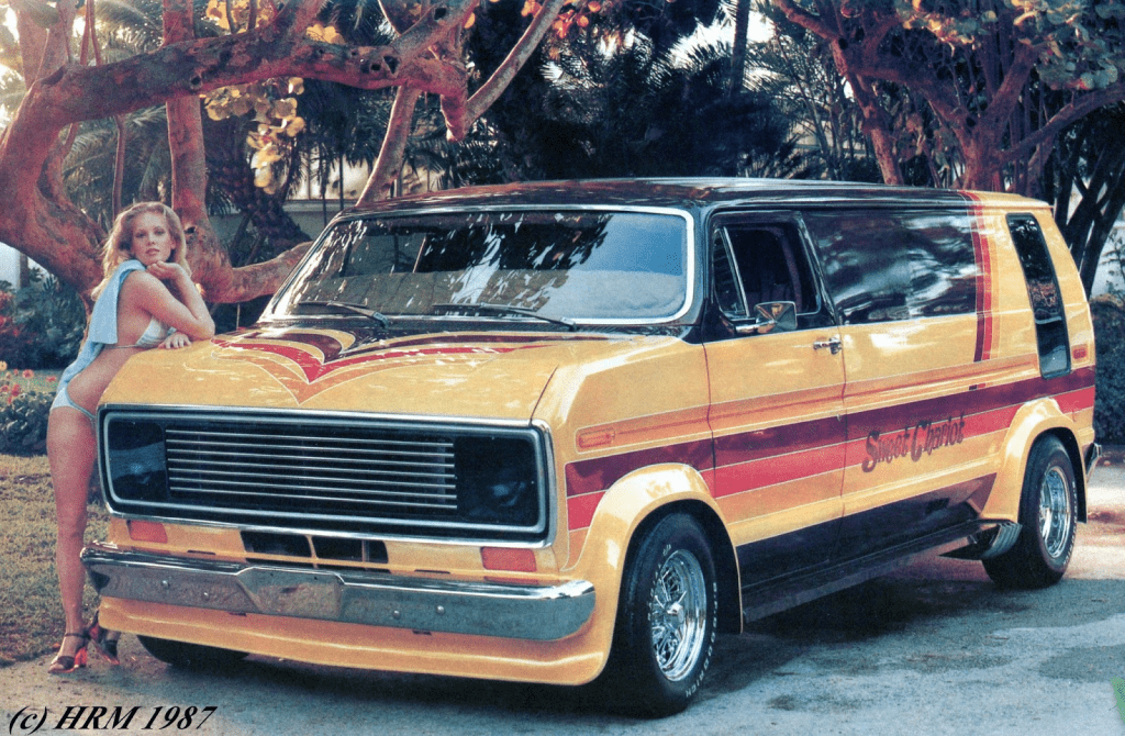 Vintage 1987 customized van Sweet Chariot with a person in swimwear leaning against it under a tree.