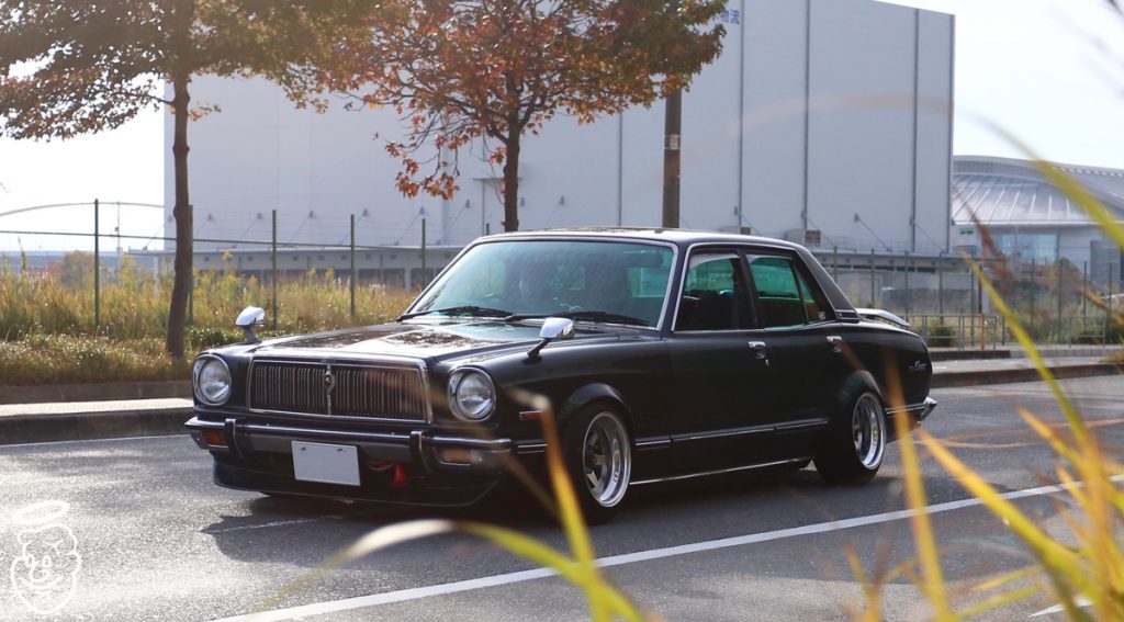 Classic black car driving on a city street with autumn trees and buildings in the background. Japanese license plate visible.