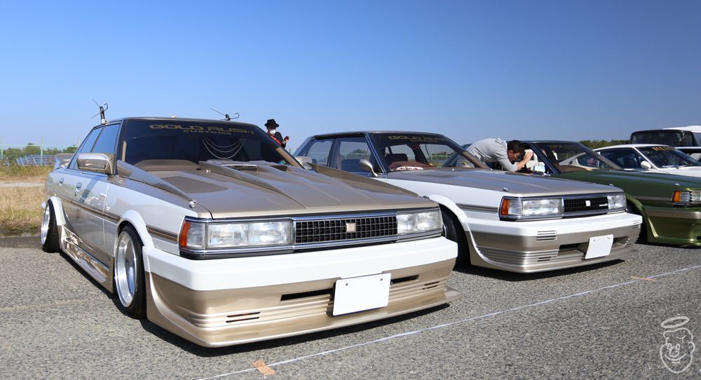 Vintage Japanese cars with custom modifications at a car show, featuring gold and white paint jobs and prominent front grills.