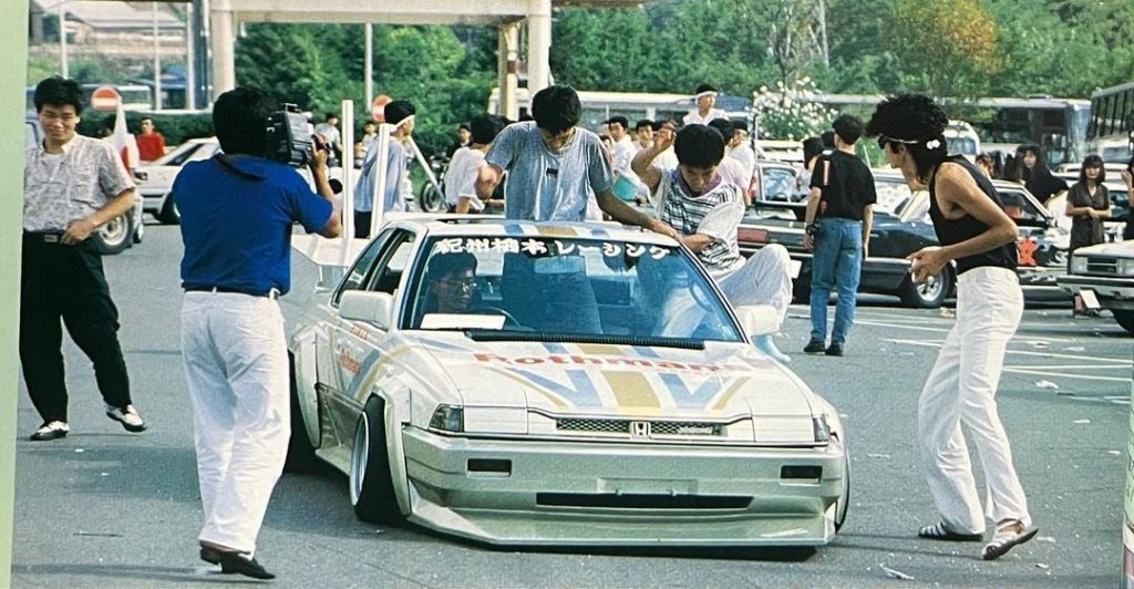 Japanese street racing crew with modified Honda and camera crew at event.