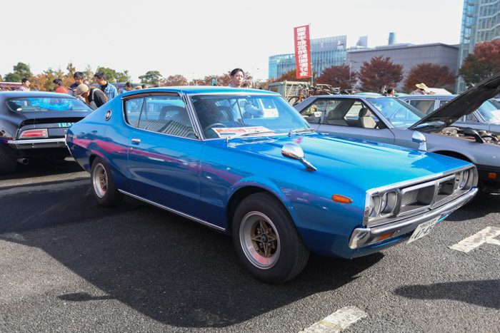 A blue classic Japanese car showcased at an outdoor car event. Japanese sign in the background translates to Historic Automobile.