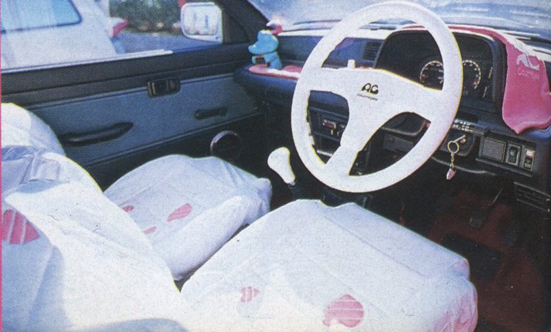 Vintage car interior with white AC steering wheel cover and pink-themed seat covers, Japanese keychain hanging on dashboard.