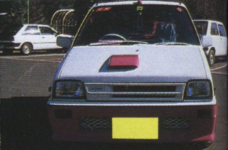 Front view of a modified white and red car with a yellow plate, parked in a lot next to other white cars.
