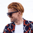 Fashionable man with red hair and sunglasses smiling, wearing a patterned shirt.