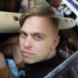 A person with blonde hair and blue eyes sitting in a vehicle, looking at the camera with a serious expression.