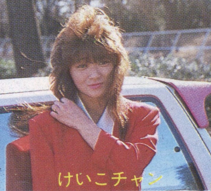 Woman in red attire standing by a car, outdoors, with the text Keiko-chan in Japanese at the bottom of the image.