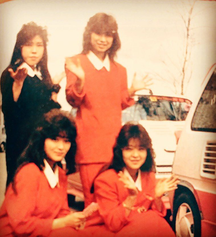 Four women in red uniforms posing cheerfully in front of a car, with trees and a clear sky in the background.
