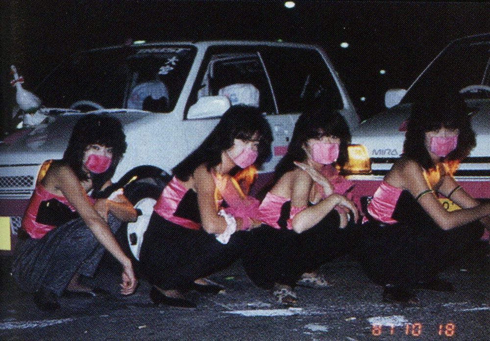Four women wearing matching outfits and pink masks squat in front of cars at night in Japan.