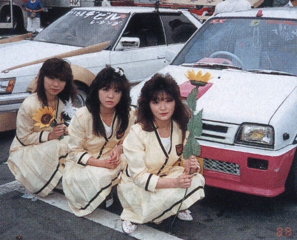 Three women in uniforms holding flowers in front of decorated cars in a Japanese car show. レディース on car windshield.Explanation:
The alt text describes the main elements in the image including the women, their uniforms, the flowers they are holding, and the Japanese car show setting to ensure it is relevant and improves SEO.