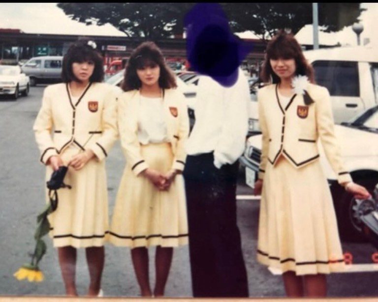 Three young women in yellow uniforms standing outdoors with blurred person, holding flowers, in front of parked cars and trees.