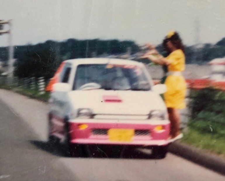 A woman in a yellow outfit poses by a white and pink car on a scenic road in Japan, conveying a lively and colorful scene.
