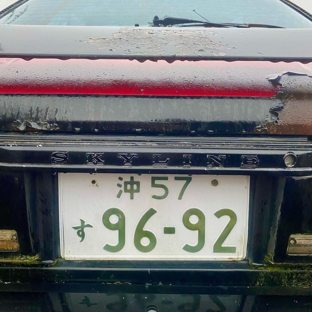 Close-up of a rusty black Nissan Skyline with Okinawa license plate 57-96-92. Text SKYLINE visible on car.