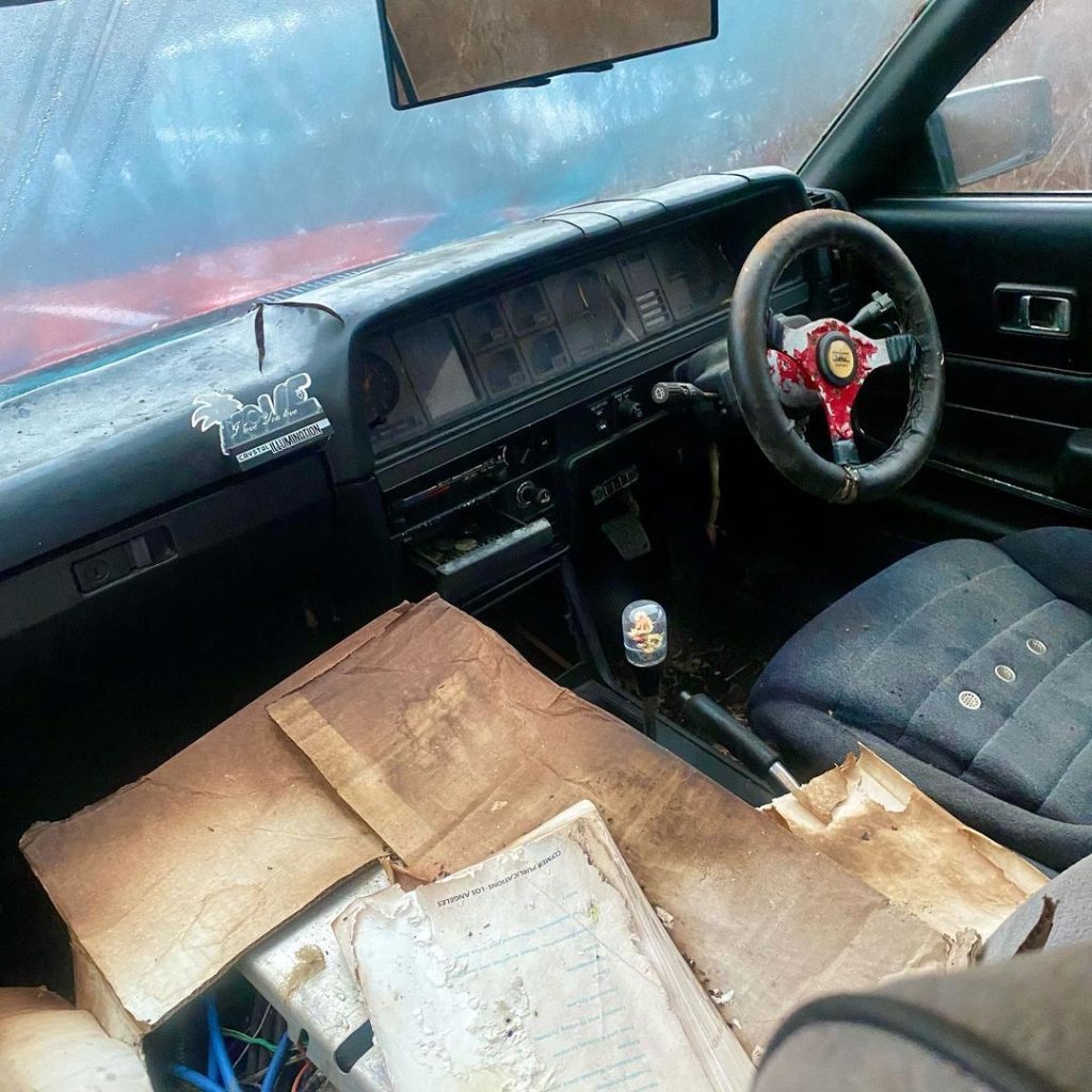 Abandoned car interior with a worn-out steering wheel and cluttered dashboard, featuring a sticker reading PEACE SPORTS PROMOTION.
