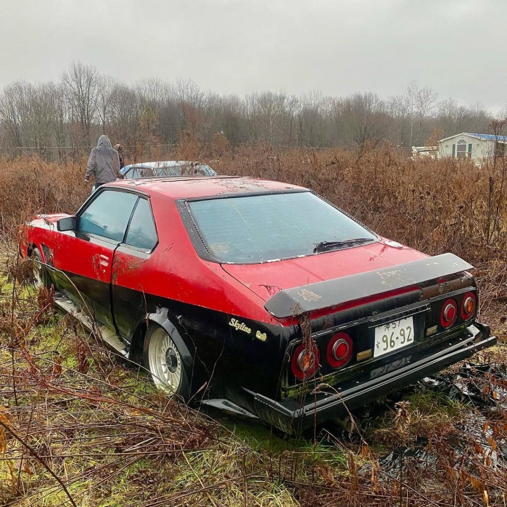 Abandoned vintage red and black Nissan Skyline car with Japanese license plate in overgrown field.