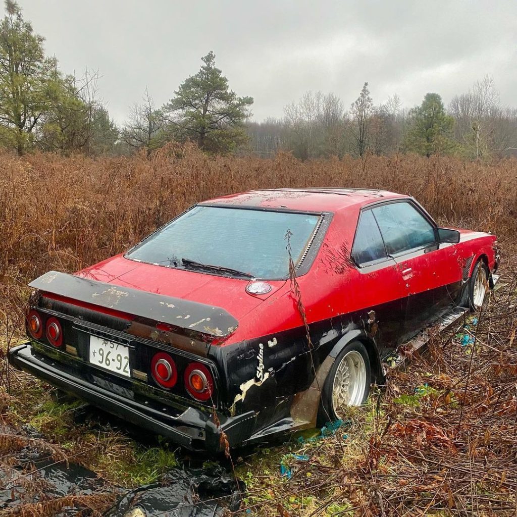 Abandoned red and black Nissan Skyline with Japanese license plate in a field of tall grass, trees in the background.