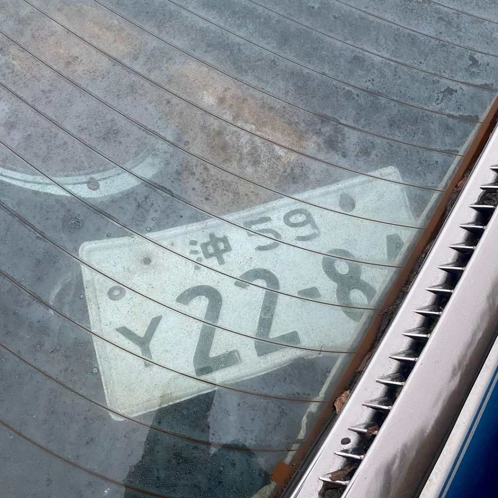 A dusty Japanese license plate with numbers 59-22-84 seen through a car's rear window, showing the kanji 沖 (Okinawa).