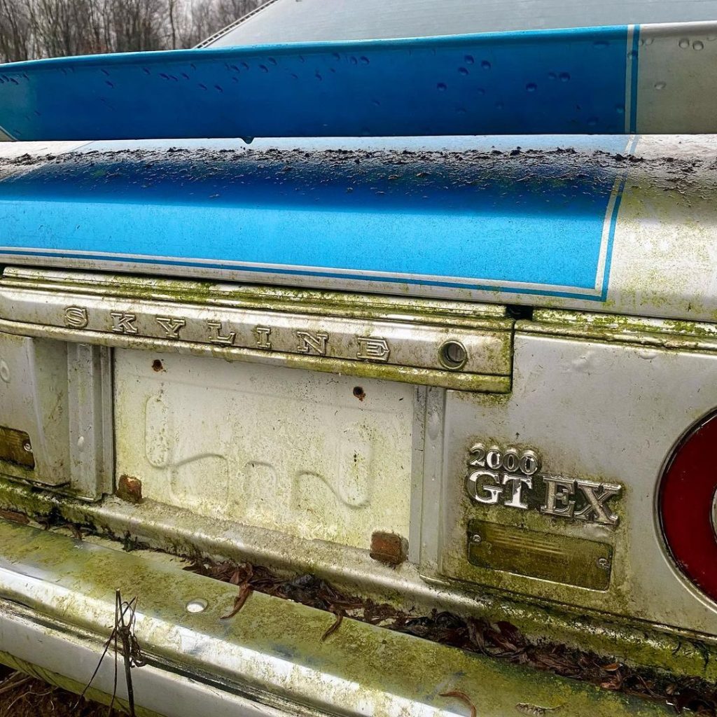 Rear view of an old and moss-covered Nissan Skyline 2000 GT-EX with a blue spoiler in a forested area.