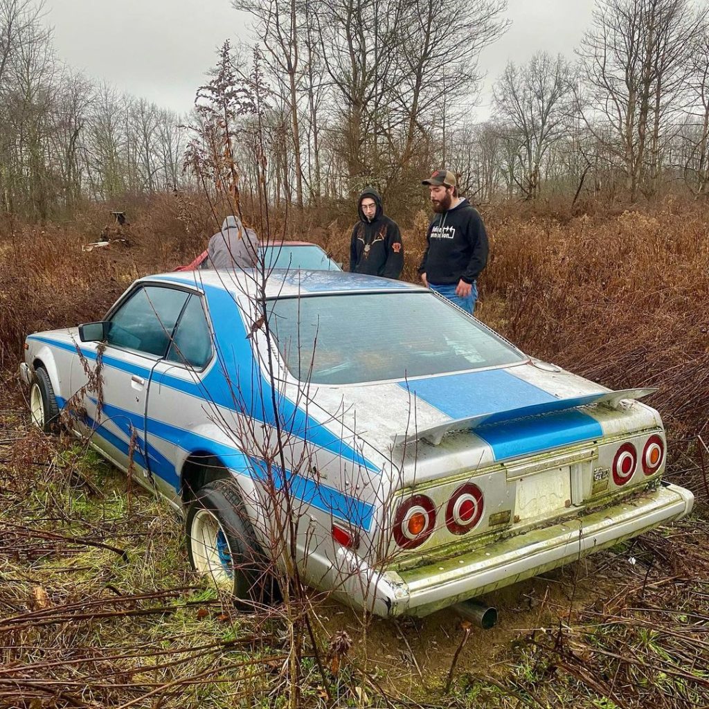 Vintage Nissan Skyline in an overgrown field with three individuals inspecting it. They appear to discuss or assess the car.
