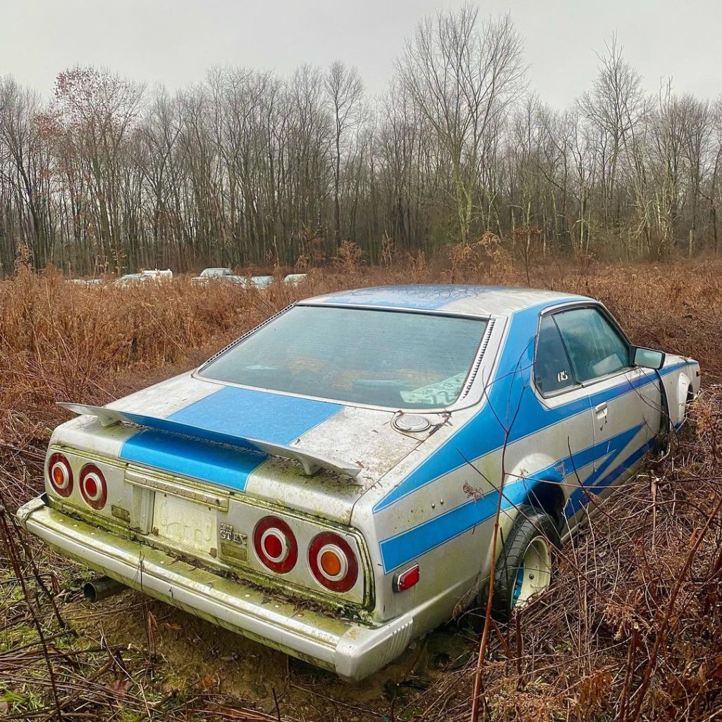 Abandoned vintage blue and white car with Japanese license plate in a field during autumn, surrounded by bare trees.