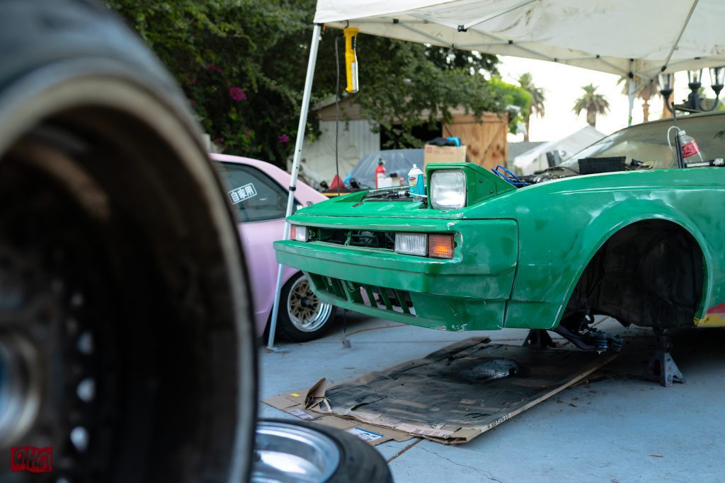 Green car being serviced under a white tent beside a pink car with Japanese text at outdoor auto repair workshop.