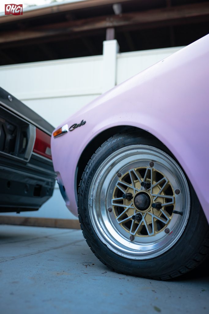 Close-up of a vintage car's front wheel with intricate gold alloy rim design and Okai emblem on the pink fender.