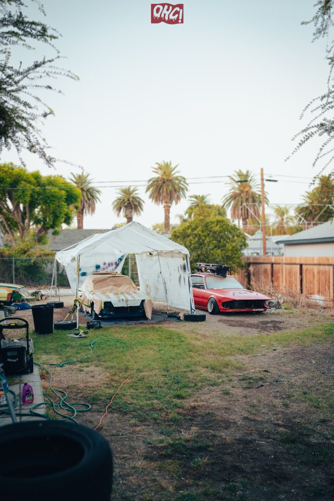 Backyard car repair and restoration scene with several cars, a plastic canopy, palm trees, and equipment under a clear sky.