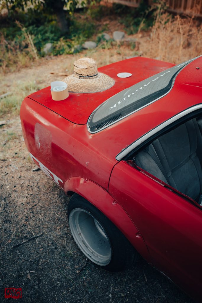 A classic red car with a wide tire, straw hat, and tape on its trunk, parked outdoors in a grassy area.