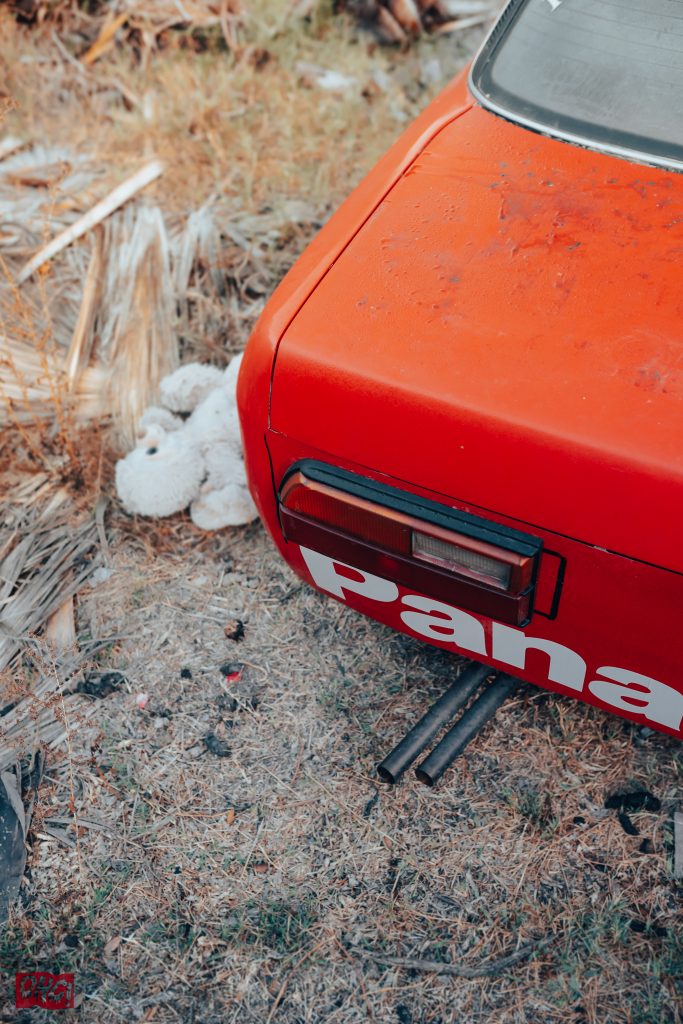 Alt text: Rear view of an orange vintage car with 'Pan' text and a stuffed toy on the ground beside it.