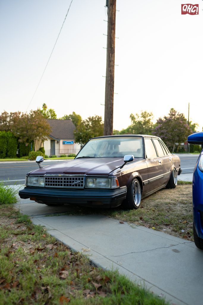 Vintage Toyota Crown sedan parked on the grass beside a residential sidewalk, with the オオクジラ!” (Ohki) logo in the background.