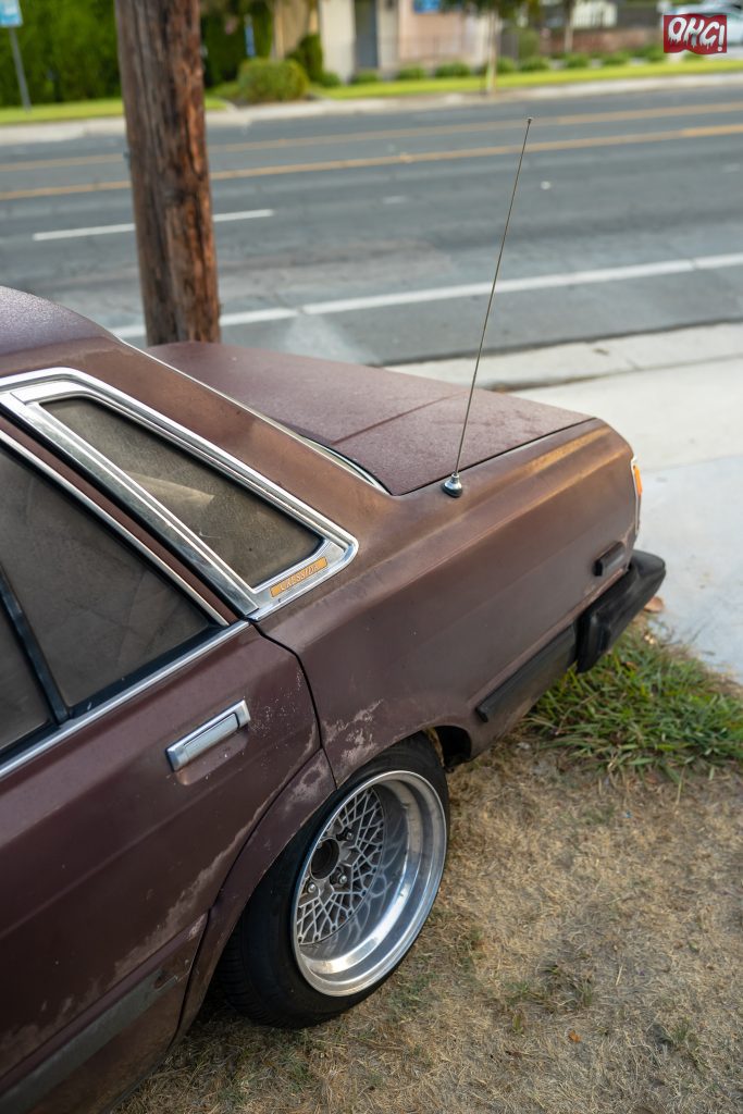 Rusty brown vintage car with custom alloy wheels parked beside a road, antenna extended.