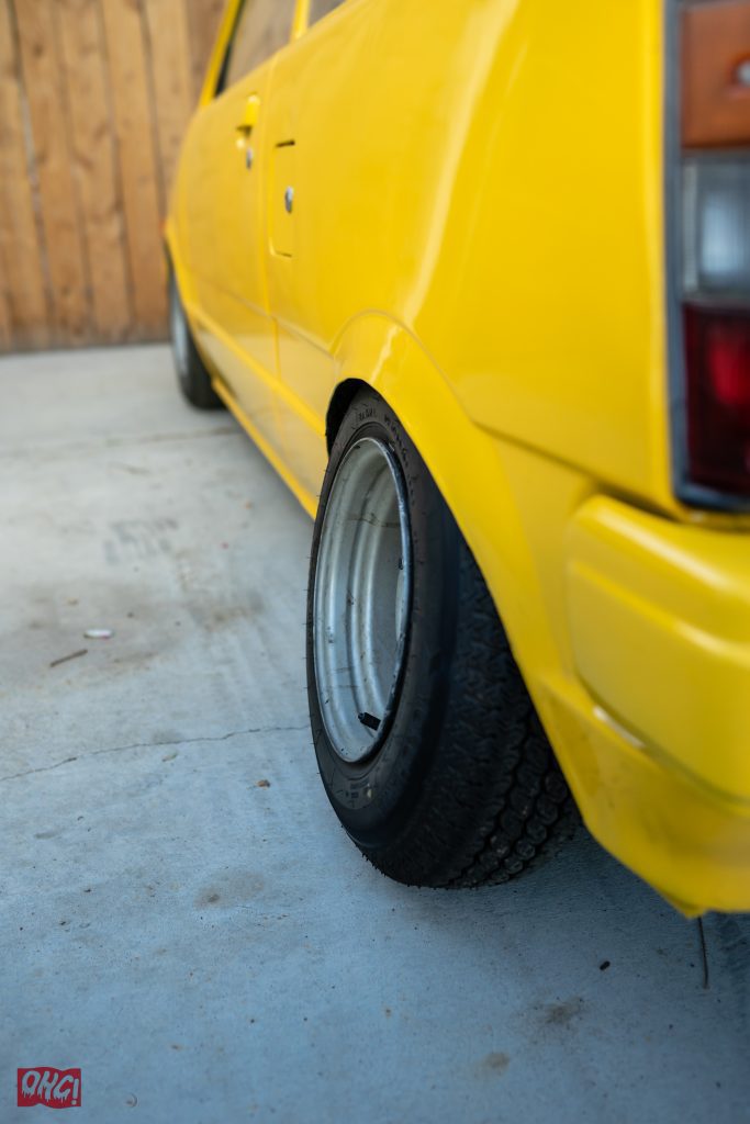 Close-up of a modified yellow car's wheel and fender, showing its wide tire stance. Japanese text reads OMG! in red.