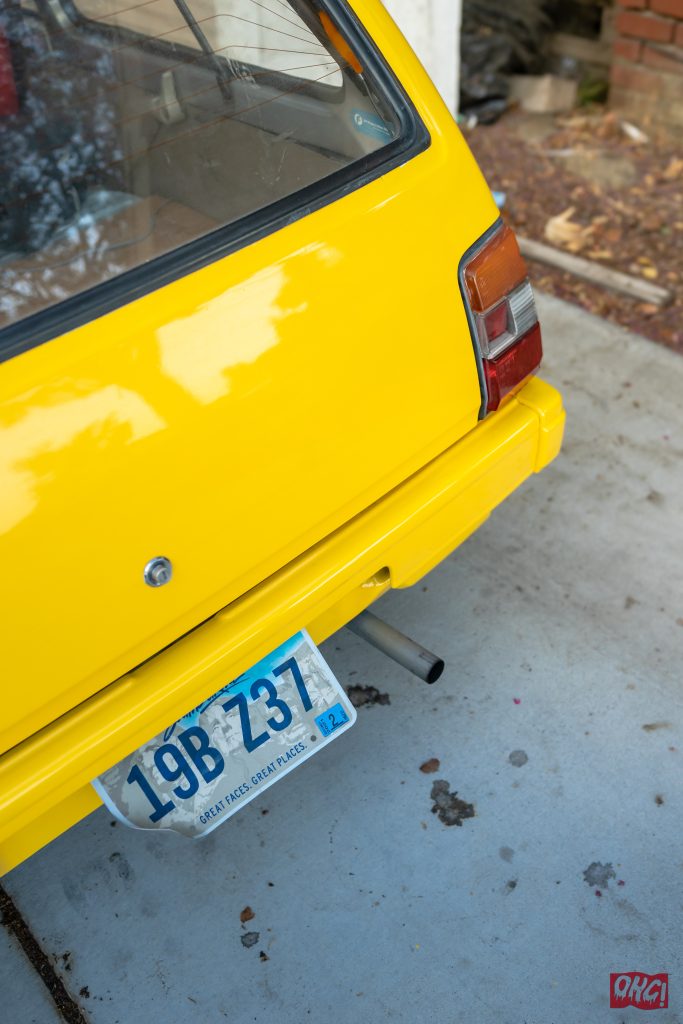Rear view of a yellow car with a blue license plate reading 19B Z37, parked on a cement surface. OKG logo in corner.