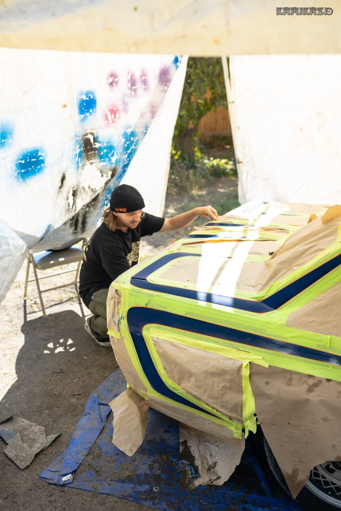Man painting a car hood with masking tape and patterns in a tarp-covered outdoor space. Japanese text reads カミカゼ (Kamikaze).