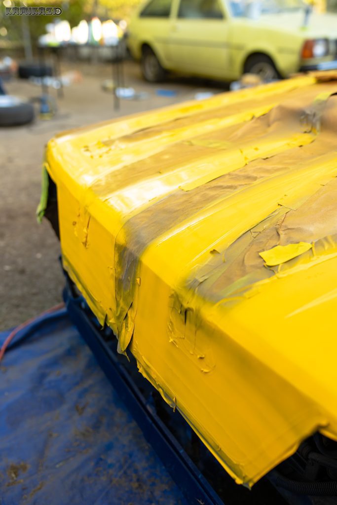 Yellow car panel being painted with masking tape on edge, tools, and another yellow car in the background.
